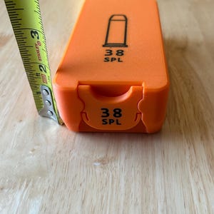 K&ouml;nnte beinhalten: Hellorangefarbene Munitionskiste mit einer schwarzen Geschossgrafik und dem Text "38 SPL". Die Kiste ist etwa 7,6 cm lang, gemessen mit einem gelb-schwarzen Ma&szlig;band.