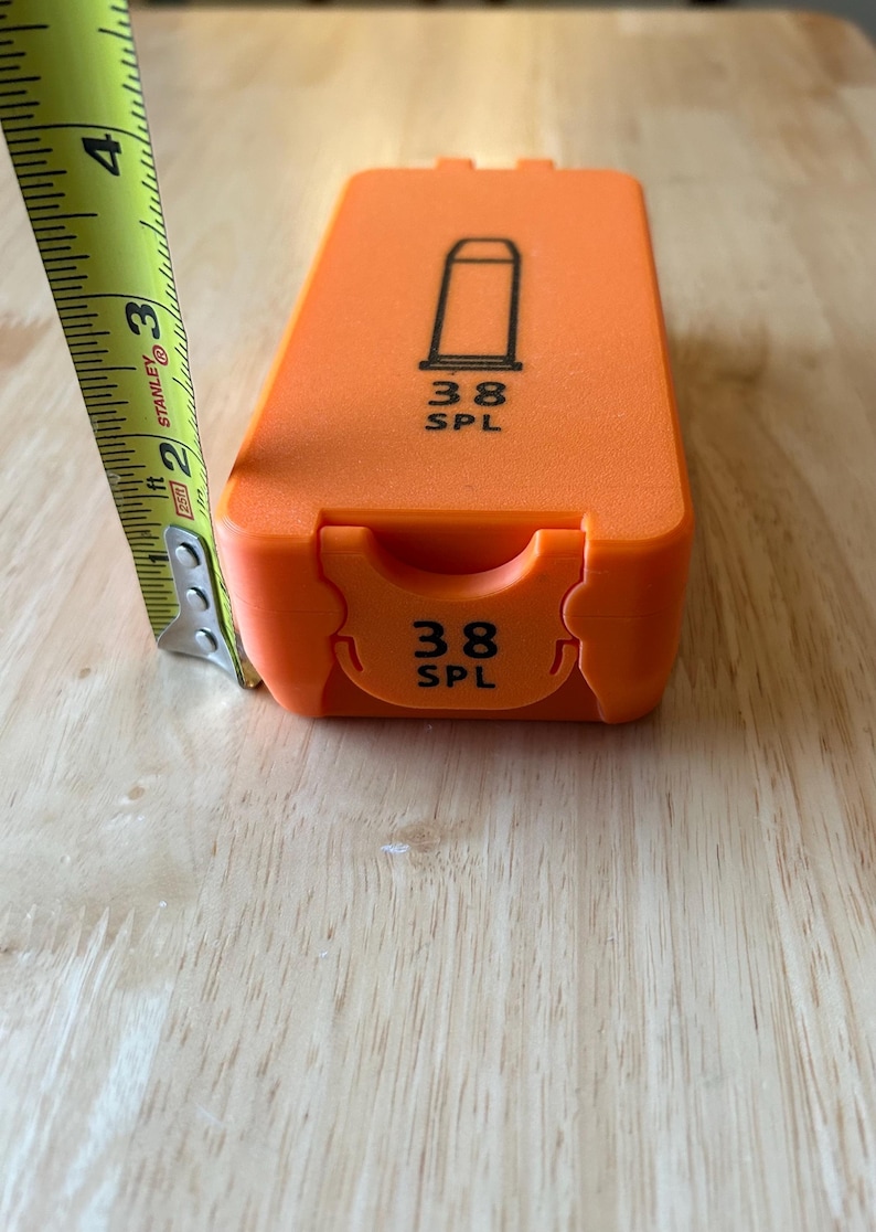 K&ouml;nnte beinhalten: Hellorangefarbene Munitionskiste mit einer schwarzen Geschossgrafik und dem Text "38 SPL". Die Kiste ist etwa 7,6 cm lang, gemessen mit einem gelb-schwarzen Ma&szlig;band.