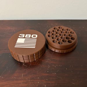 May include: A brown, 3D-printed container with a lid featuring the number "380" and a white graphic. The container is open, revealing a grid of small holes. The design is likely for organizing small items.