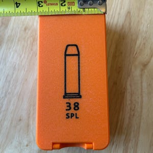 K&ouml;nnte beinhalten: Hellorangefarbene rechteckige H&uuml;lle mit schwarzer Umrisszeichnung einer Patrone und dem Text "38 SPL". Die H&uuml;lle wird mit einem gelben Ma&szlig;band gemessen, die Breite betr&auml;gt ca. 5 cm.