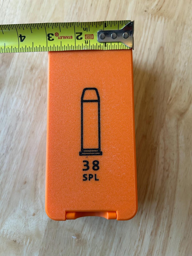 K&ouml;nnte beinhalten: Hellorangefarbene rechteckige H&uuml;lle mit schwarzer Umrisszeichnung einer Patrone und dem Text "38 SPL". Die H&uuml;lle wird mit einem gelben Ma&szlig;band gemessen, die Breite betr&auml;gt ca. 5 cm.