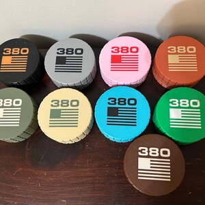 May include: A collection of round, colorful bottle stoppers. Each stopper features the number "380" above a horizontal striped design, resembling a flag. Colors include black, gray, pink, orange, brown, green, blue, and tan.