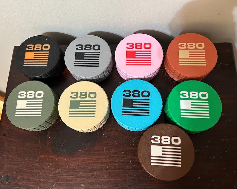 May include: A collection of round, colorful bottle stoppers. Each stopper features the number "380" above a horizontal striped design, resembling a flag. Colors include black, gray, pink, orange, brown, green, blue, and tan.