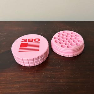 May include: Pink, cylindrical container with the number "380" and a red and white flag design on the lid. The container is open, revealing a grid of small holes. The container is made of plastic.