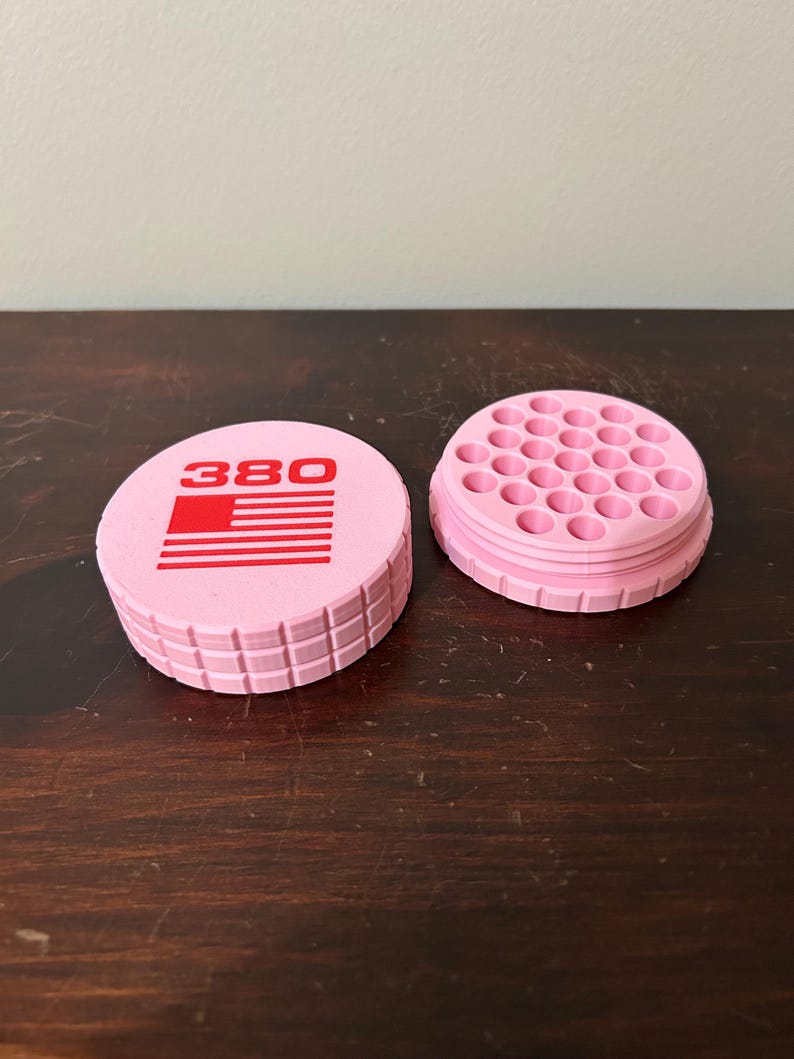 May include: Pink, cylindrical container with the number "380" and a red and white flag design on the lid. The container is open, revealing a grid of small holes. The container is made of plastic.