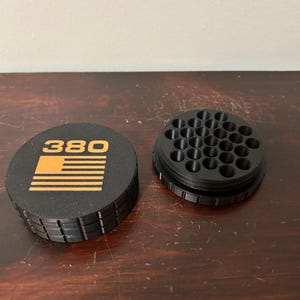 May include: Black and orange container with the number "380" and a flag design. The container is round with a ridged edge. The lid is separate from the base, which has multiple holes.