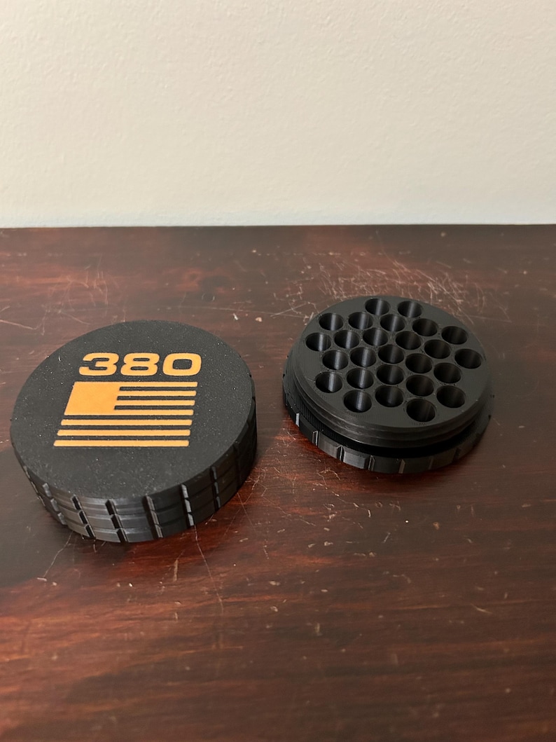 May include: Black and orange container with the number "380" and a flag design. The container is round with a ridged edge. The lid is separate from the base, which has multiple holes.