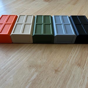 May include: Five rectangular boxes in a row, coloured orange, cream, olive green, light grey, and black. Each box features a rectangular grid design on the top. The boxes are arranged on a light wooden surface. Measurements are in centimetres.