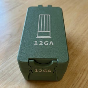 Könnte beinhalten: Ein olivgrüner rechteckiger Behälter mit dem weißen Aufdruck "12GA". Die Oberseite des Behälters hat eine Grafik mit vertikalen Linien. Der Behälter steht auf einer Holzoberfläche.