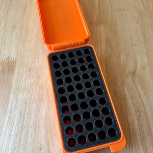 K&ouml;nnte beinhalten: Eine offene, leuchtend orangefarbene Kunststoff-Aufbewahrungsbox mit einem schwarzen Schaumstoffeinsatz. Der Schaumstoff hat mehrere kreisf&ouml;rmige Aussparungen. Die Box ist rechteckig mit einem Klappdeckel und liegt auf einer hellen Holzoberfl&auml;che.