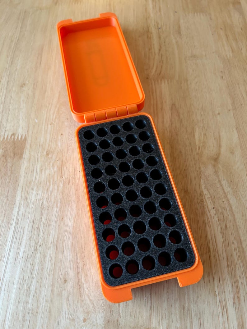 K&ouml;nnte beinhalten: Eine offene, leuchtend orangefarbene Kunststoff-Aufbewahrungsbox mit einem schwarzen Schaumstoffeinsatz. Der Schaumstoff hat mehrere kreisf&ouml;rmige Aussparungen. Die Box ist rechteckig mit einem Klappdeckel und liegt auf einer hellen Holzoberfl&auml;che.