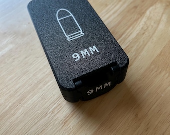 May include: Black rectangular case with a white bullet graphic and the text "9MM". The case is hinged and appears to be made of a textured material. The case is on a wooden surface.