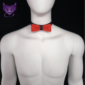 May include: A red bow tie choker necklace with a black leather strap. The choker is worn on a white mannequin.