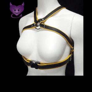 May include: Black and yellow leather harness with a metal ring and buckle closure. The harness features yellow stitching and trim, and is displayed on a white mannequin against a black background. A purple devil heart graphic is in the upper left corner.