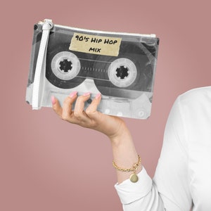 Puede incluir: Una cartera de mano blanca con forma de casete con un panel frontal de plástico transparente. El panel frontal presenta una imagen en blanco y negro de un casete con el texto "90's Hip Hop Mix" en una etiqueta marrón.