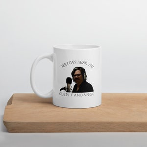 May include: White ceramic mug with a black and white image of a man with glasses holding a microphone. The text "YES, I CAN HEAR YOU" is above the image and "CLEM FANDANGO" is below the image.