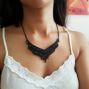 Puede incluir: Un collar de macramé negro con un diseño en forma de V. El collar presenta pequeñas cuentas oscuras y un delicado patrón tejido. El collar se usa sobre una camiseta sin mangas blanca.