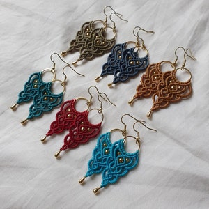 May include: Five pairs of handmade earrings with intricate knotted designs. The earrings are in various colors, including teal, brown, red, and blue. Each earring features a gold hoop and a gold bead accent.