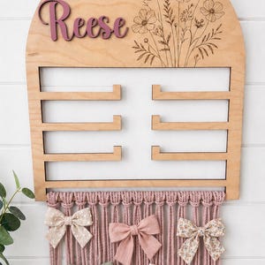 Puede incluir: Un organizador de pared de madera con parte superior redondeada, que presenta el nombre Reese en letra cursiva morada y un diseño floral grabado con láser. Incluye tres barras horizontales de almacenamiento y flecos de macramé rosa con cinco lazos.