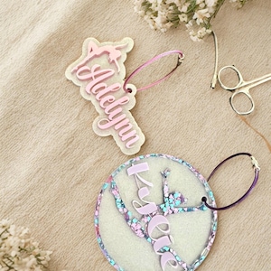 May include: Two personalised acrylic keyrings. One is shaped like a gymnast with the name "Adelynn" in pink script. The other is a circle with a gymnast design and the name "Kylie" in pink. Both have matching coloured loops.