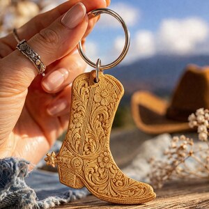 May include: A tan-colored cowboy boot keychain with intricate floral and scrollwork details. The boot features a spur and is attached to a silver keyring. The background includes a blurred view of a cowboy hat and a mountain range.