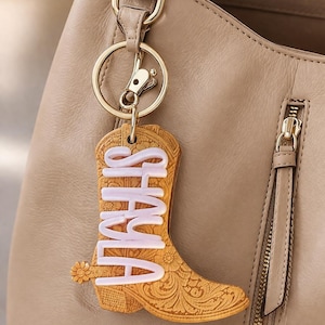May include: A tan cowboy boot keychain with the name "SHALA" in white, hanging from a gold-colored keyring. The boot has a floral design and a small flower detail. The background includes a tan handbag.