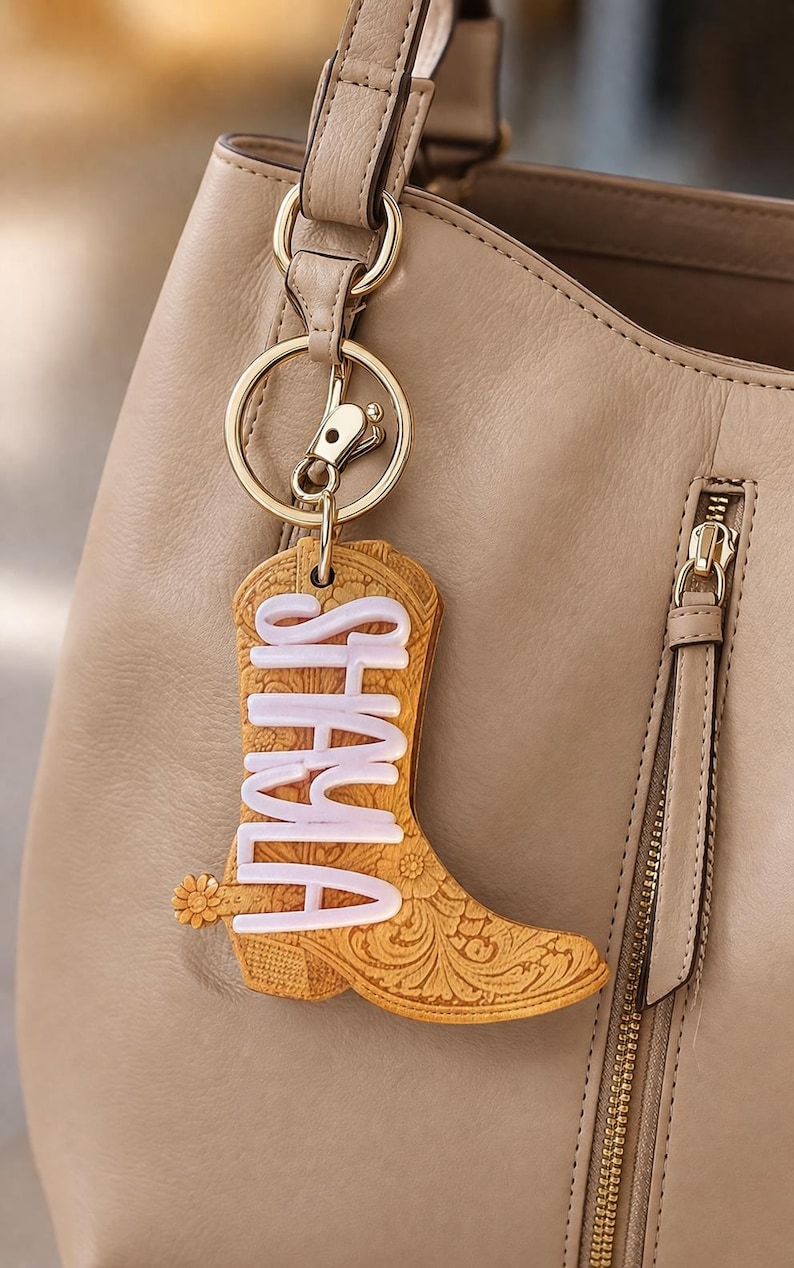 May include: A tan cowboy boot keychain with the name "SHALA" in white, hanging from a gold-colored keyring. The boot has a floral design and a small flower detail. The background includes a tan handbag.