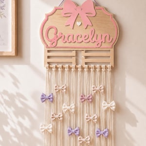 May include: A wooden wall-mounted organizer featuring a pink bow at the top and the name Gracelyn in pink script. Below the wooden plaque, multiple long cream-colored macrame cords hang down, each decorated with small fabric bows in various colors.