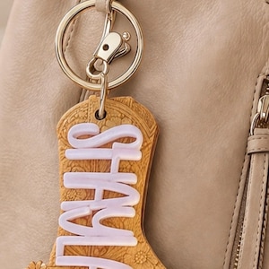 May include: A tan leather keychain in the shape of a cowboy boot with the name "SHALA" in white lettering. The boot has floral and scrollwork details and is attached to a gold-colored keyring and clasp.
