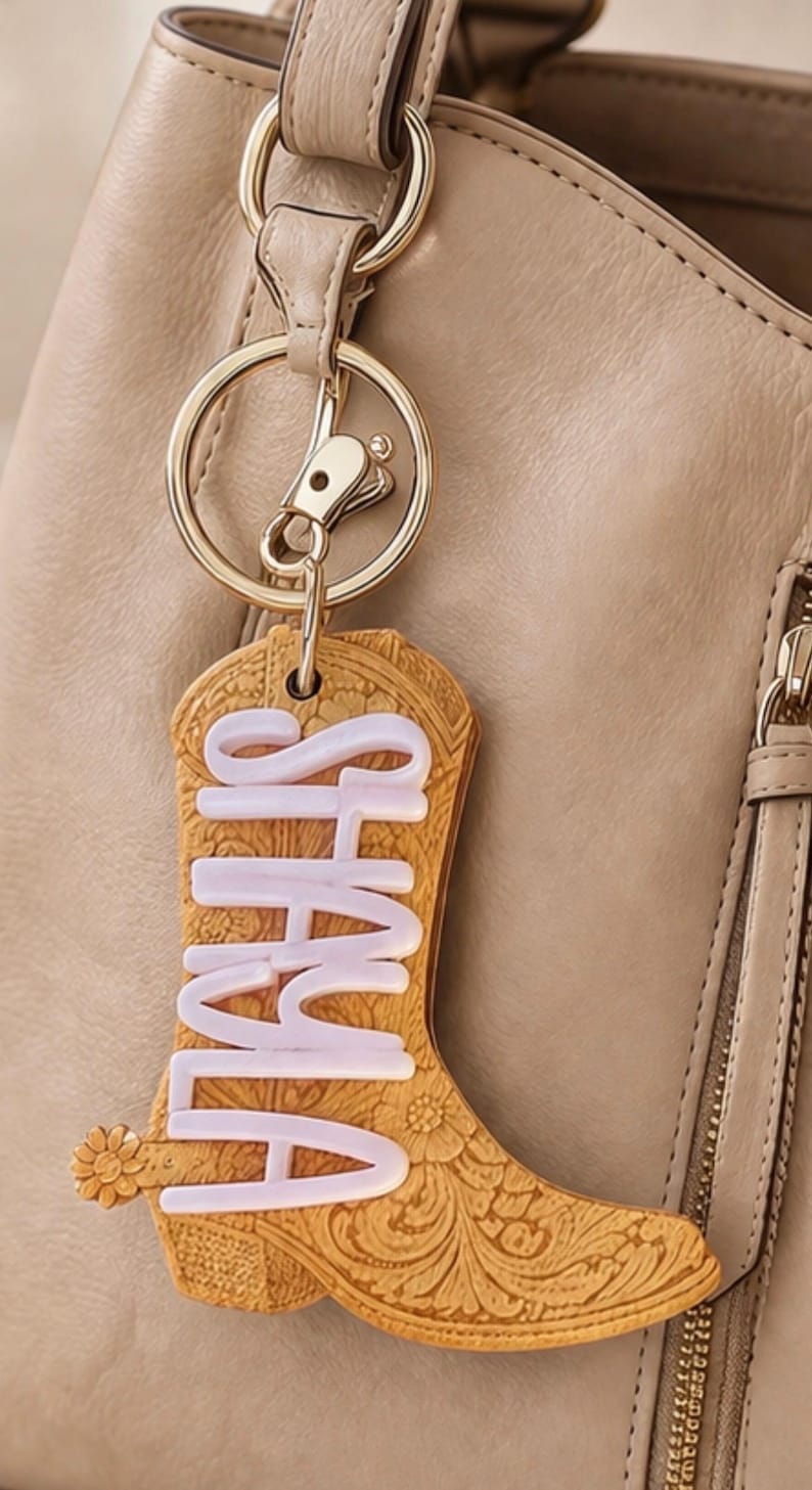 May include: A tan leather keychain in the shape of a cowboy boot with the name "SHALA" in white lettering. The boot has floral and scrollwork details and is attached to a gold-colored keyring and clasp.