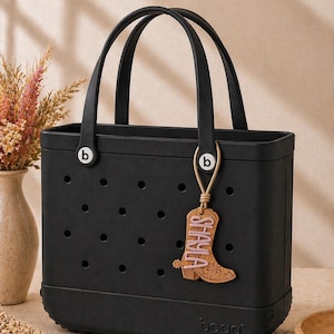 May include: Black tote bag with two handles and a decorative charm. The charm is a boot shape with the name "SHAYLA" in pink lettering. The bag has the brand name "bogg" on the bottom.