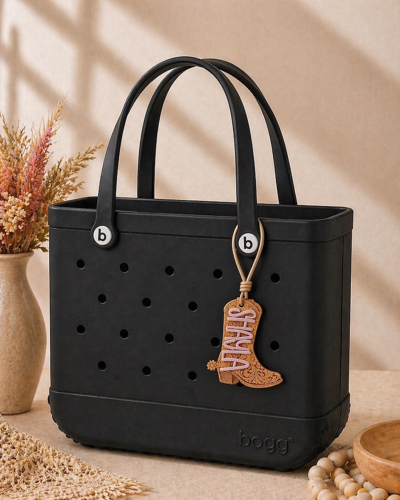 May include: Black tote bag with two handles and a decorative charm. The charm is a boot shape with the name "SHAYLA" in pink lettering. The bag has the brand name "bogg" on the bottom.
