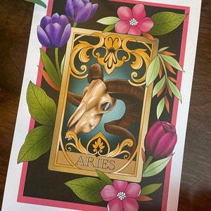 May include: A tarot card with a gold frame and ornate details. The card features a ram skull with a blue background and the word "ARIES" in gold lettering. The card is surrounded by purple and pink flowers and green leaves.