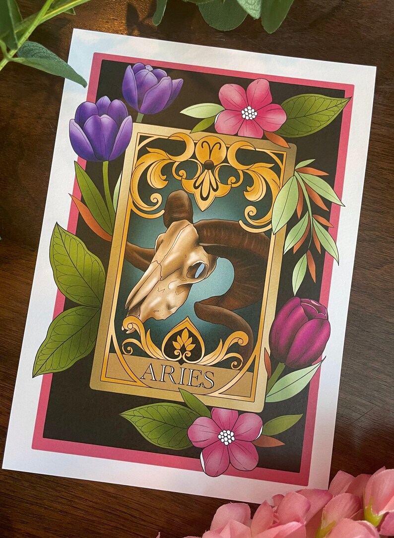 May include: A tarot card with a gold frame and ornate details. The card features a ram skull with a blue background and the word "ARIES" in gold lettering. The card is surrounded by purple and pink flowers and green leaves.
