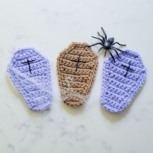 May include: Three crocheted coffin-shaped ornaments, two are purple and one is brown, with a black plastic spider on a white background.
