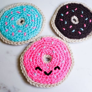 Könnte beinhalten: Drei gehäkelte Donuts, einer rosa mit einem lächelnden Gesicht, einer blau mit weißen Streuseln und einer braun mit rosa und blauen Streuseln.