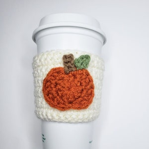 May include: White crocheted coffee cup cozy with an orange pumpkin design. The pumpkin has a brown stem and green leaves.