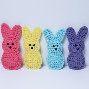 Four small crocheted peeps sit in a row along a white background. From left to right their colors are pink, yellow, blue and purple. Each has two tiny dots for eyes and one tiny dot nose.