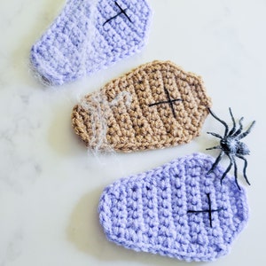 A set of three crochet coffin coasters in a row with spider webs and a spider on a marble countertop.