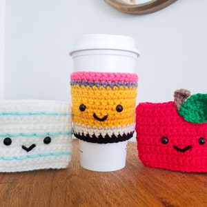 On a wooden table top from left to right is a piece of paper crochet cozy, a pencil and an apple. Each cozy is hand crocheted with yarn and has a smiling little face. The pencil cozy is on a white mug and the others are standing alone.