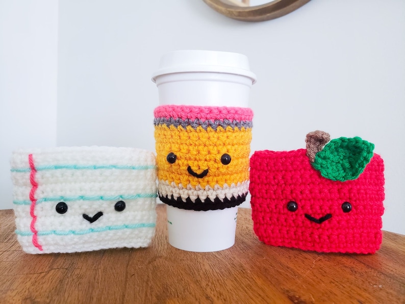 On a wooden table top from left to right is a piece of paper crochet cozy, a pencil and an apple. Each cozy is hand crocheted with yarn and has a smiling little face. The pencil cozy is on a white mug and the others are standing alone.