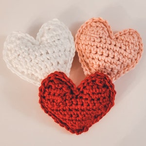 A set of three large crochet heart plushies made of bulky / chunky yarn against a white backdrop. There is one pink, one red, and one white heart.