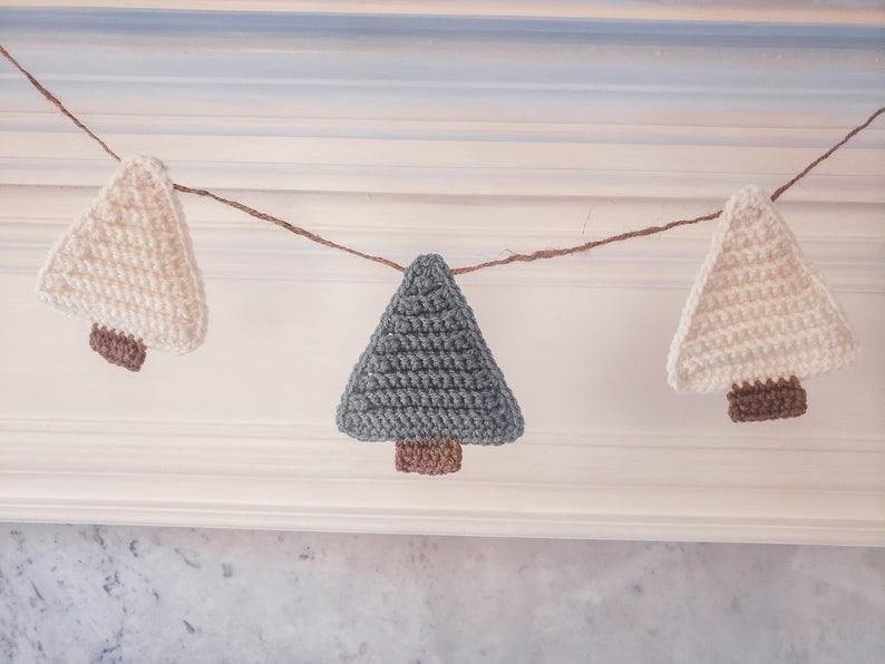 Crocheted Christmas Trees strung into a garland draped across a white mantle with jute. The trees are made of green and cream yarn with light brown trunks.