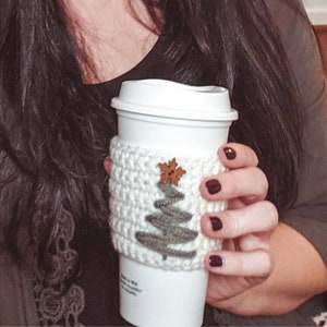 The crochet Christmas Tree Cozy in use on a white Starbucks travel mug being held by a hand with painted nails.