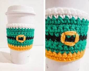 May include: A green, black, and yellow crocheted coffee cup cozy with a white top. The cozy has a black belt with a gold buckle.