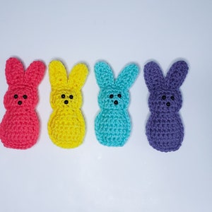 Four small crocheted peeps sit in a row along a white background. From left to right their colors are pink, yellow, blue and purple. Each has two tiny dots for eyes and one tiny dot nose.