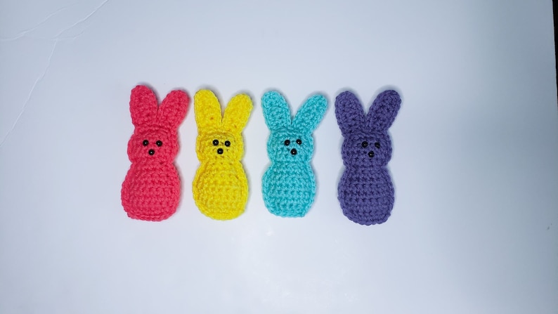 Four small crocheted peeps sit in a row along a white background. From left to right their colors are pink, yellow, blue and purple. Each has two tiny dots for eyes and one tiny dot nose.