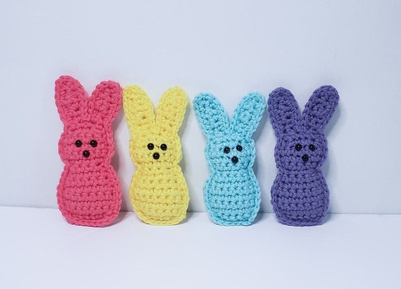 Four small crocheted peeps sit in a row along a white background. From left to right their colors are pink, yellow, blue and purple. Each has two tiny dots for eyes and one tiny dot nose.