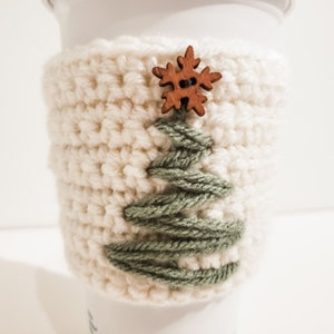 An up close image of the crocheted Christmas Tree coffee cozy displayed on a white Starbucks mug with a white background. The cup cozy is minimalist using green and cream yarn and a wooden button.