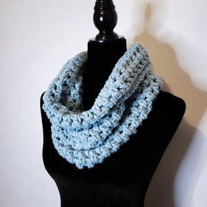 A light blue crochet fleece cowl displayed on a black velvet mannequin against a white backdrop at a side angle.